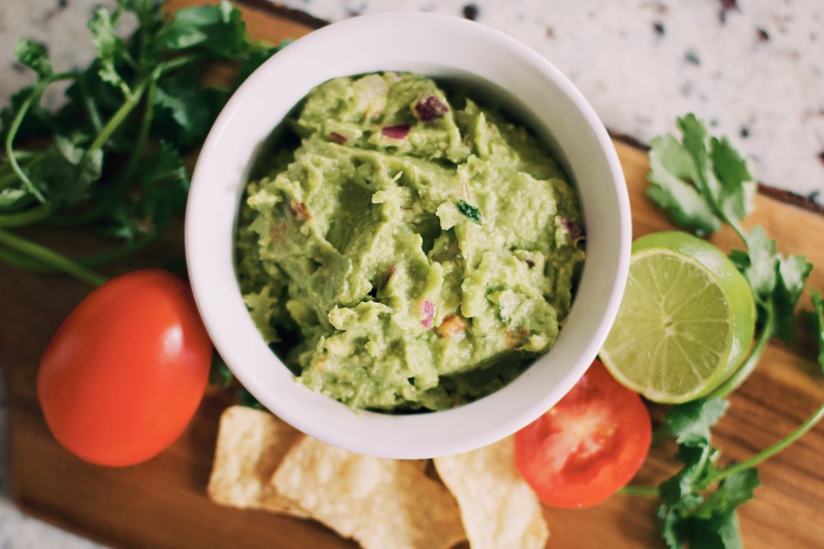 Guacamole in a white bowl surrounded by limes, tomatoes, and cilantro