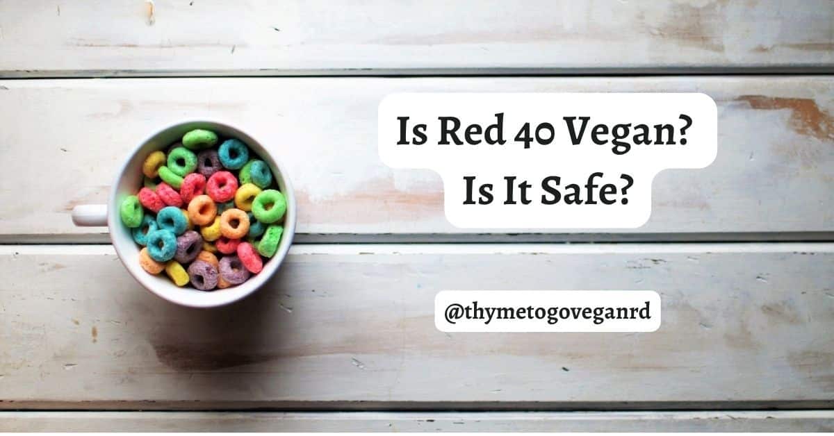 Brightly colored cereal in a white bowl on a gray wooden table with text overlay reading "is red 40 vegan? is it safe? @thymetogoveganRD"
